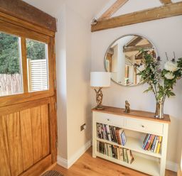 A hallway with a wooden door a white console table with books flowers and a lamp below a round mirror at The Old Piggery in Defford