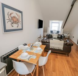 A dining area with a wooden table and white chairs next to a living room with brown leather sofas and a wall-mounted television at Foxton Penthouse in Whitby