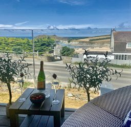 A balcony with a wooden table holding a bottle of champagne and two glasses overlooking a beach and road at Porth Beach in Porth near St Columb Minor