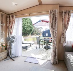 A living room with a sofa TV fan and sliding door opening to a patio with table and chairs at Freddys Lodge Foxburrow Hang in Fritton