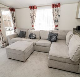 A living room with a gray sectional sofa, round and square cushions, floral curtains, and a sliding glass door at Moonlight Lodge in Bockenfield Country Holiday Park near West Thirston