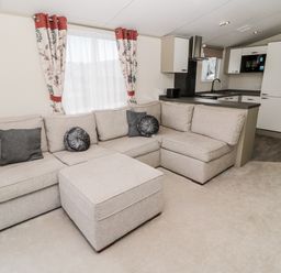 A living room with a beige sectional sofa and ottoman adjacent to a kitchen with white cabinets at Moonlight Lodge in Bockenfield Country Holiday Park near West Thirston