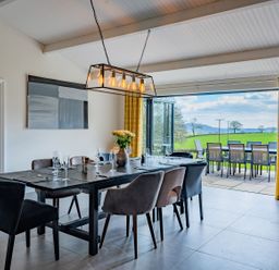 A dining room with a black table and mixed chairs next to open doors leading to a patio with outdoor chairs and tables at Quantock Barn in Stockland near Honiton