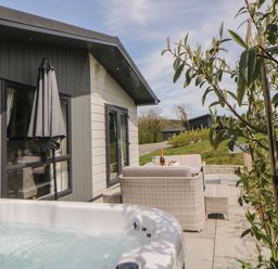 An outdoor patio area with a hot tub a table with chairs and a bottle of wine next to a modern house at Meadow Lodge in Lifton