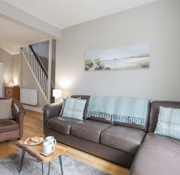 A living room with a brown leather sofa and armchair a wooden table with cups and croissants and a painting on the wall at Cobblers Cottage in Beaumaris