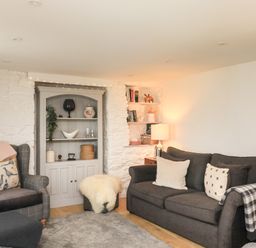 A living room with a dark sofa and gray armchair next to a white stone wall with shelves and a lamp at Hope Cottage in Buckfastleigh