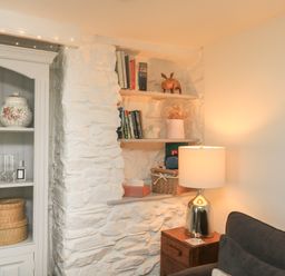 A corner with white stone walls featuring built-in shelves with books and decorative items a wooden side table with a lamp and a dark sofa at Hope Cottage in Buckfastleigh