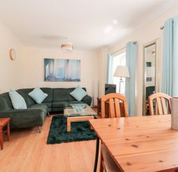A living room with a green sofa blue cushions a glass coffee table wooden dining table and chairs near windows with blue curtains at Garland in Nottington near Weymouth