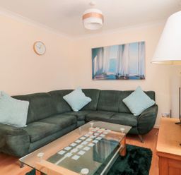 A living room with a dark green sectional sofa and light blue cushions a glass coffee table a wooden side table with a lamp a wall clock and a wall picture at Garland in Nottington near Weymouth