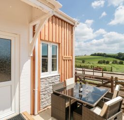 An outdoor patio area with a glass table and four wicker chairs next to a wooden building with a window and a sign reading Emminster with green fields and a wooden fence in the background at Emminster in Nottington near Weymouth