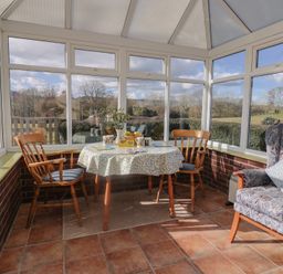 A conservatory with a round table covered with a floral tablecloth and two wooden chairs next to a patterned armchair at Broadstones in Bridport