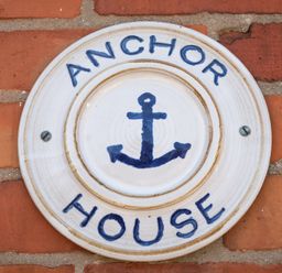 A round ceramic sign mounted on a brick wall with an anchor symbol and the words Anchor House in Weymouth