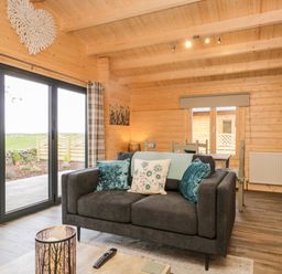 A living room with a dark grey sofa with cushions near large glass doors and a dining table in a wooden cabin at Howgill Lodge in Gatebeck near Kirkby Lonsdale