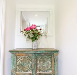 A distressed wooden cabinet with a vase of pink flowers and a white framed mirror above it at Bluebell Cottage in Mary Tavy