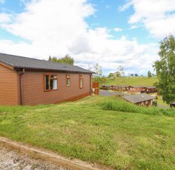 A brown wooden cabin on a grassy hill with trees and more cabins in the background at Rashierigg in Catterick North Yorkshire