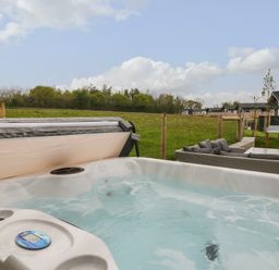 An outdoor hot tub with water and bubbles next to a patio with a sofa set in a fenced grassy area with buildings in the background at Lily in Broadwoodwidger near Ashwater
