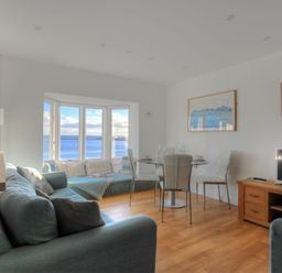 A living room with blue sofas a dining table and chairs wooden floor and a bay window with sea view at Sea Fever in Lyme Regis