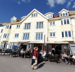 People dining outside a restaurant in front of a three-story building with white walls and multiple windows at Sea Fever in Lyme Regis