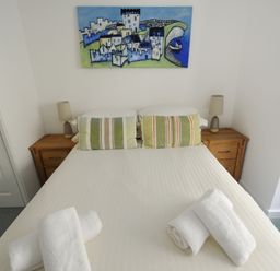 A bedroom with a double bed two wooden bedside tables lamps pillows rolled towels and a painting above the bed at Sea Fever Lyme Regis