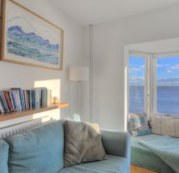 A living room with a blue sofa a shelf of books a floor lamp and a window seat with cushions overlooking the sea at Sea Fever in Lyme Regis