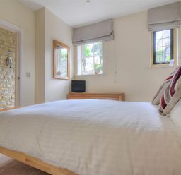 A bedroom with a wooden bed frame and white bedding a wooden dresser with a small black tv two windows with gray blinds and a stone wall visible through a doorway at Coram Lodge in Lyme Regis