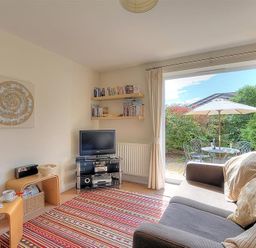 A living room with a sofa a tv on a stand shelves with books and an open door leading to a patio with a table umbrella and chairs at 1 Coppers Knapp in Lyme Regis