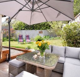 A patio with a glass table and wicker seating under a large umbrella overlooking a garden with two striped chairs at Bucket & Spade Lyme Regis