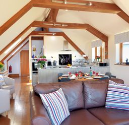 An open plan living space with a brown leather sofa and striped cushions a dining table set for six and a kitchen with wooden beams and windows at Bucket & Spade in Lyme Regis