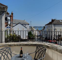 A balcony with a table and chairs overlooking a street with buildings and a view of the sea at Blacksmiths Cottage in Lyme Regis