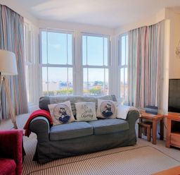A living room with a blue sofa and patterned cushions by bay windows with striped curtains at 1 Cobb View in Lyme Regis