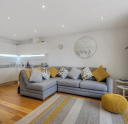 A living room with a grey sectional sofa with patterned and yellow cushions a knitted yellow pouf a patterned rug and a white kitchen with cabinets and appliances at The Lower Deck in Lyme Regis