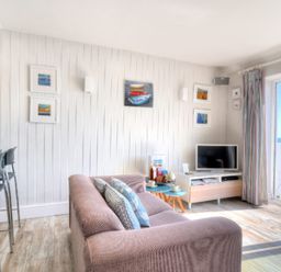 A living room with a pink sofa and pillows a small blue table a television and a balcony door showing the sea at 9 Bay View Court Lyme Regis