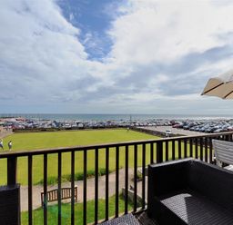 A balcony with outdoor seating and an umbrella overlooking a lawn with people and a parking area by the sea at 8 Bowling Green Chalets in Lyme Regis