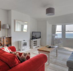 A living room with a red sofa, wooden coffee table, armchair by bay windows with ocean view, tv on cabinet and wooden sideboard at Ross House in Lyme Regis