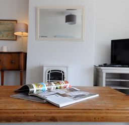 A wooden table with magazines in a living room with a lamp on a sideboard mirror above a fireplace and a television on a cabinet at Ross House in Lyme Regis