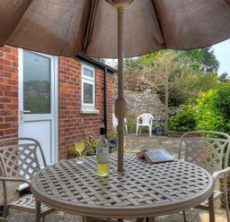 An outdoor patio with a round table under an umbrella surrounded by chairs and a brick wall with plants and flowers at 23 Lym Close Lyme Regis