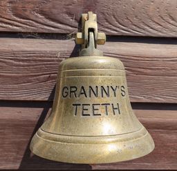 A brass bell mounted on a wooden wall with the words Granny's Teeth engraved on it at Grannys Teeth in Lyme Regis