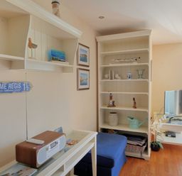 A living room with shelves holding decorative items a television on a white table and a blue ottoman at 2 Buckfields Lyme Regis