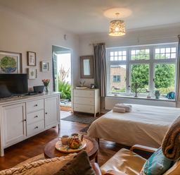 A bedroom with a bed a television on a white cabinet a round table with a teapot and cups and a window with curtains at The Apartment at Queen Anne's Lodge in Lyme Regis