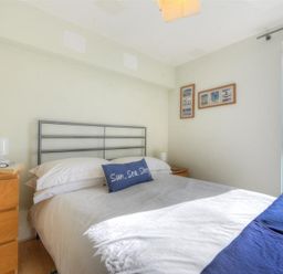 A bedroom with a bed white bedding and blue pillow beside wooden bedside tables with lamps and framed pictures on the wall at 8 Driftwood in West Bay