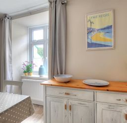 A dining area with a table covered with a polka dot cloth a window with curtains a sideboard with dishes and flowers and a Lyme Regis poster on the wall at Mermaid House in Lyme Regis