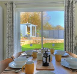 A dining table set with bowls cups and cutlery in front of sliding glass doors leading to a garden with a shed at Woody View in Colyton