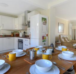 A kitchen and dining area with white cabinets yellow mugs and white bowls on a wooden table and a beige living room with leather chairs at Woody View in Colyton