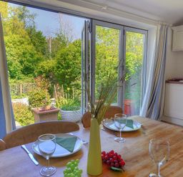A dining table set with plates glasses and cutlery beside open glass doors overlooking a garden at 24 Mill Green Lyme Regis