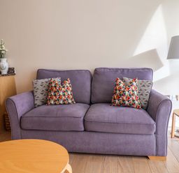 A purple sofa with patterned cushions in a room with wooden tables and a floor lamp at 2 Driftwood in West Bay