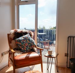 A leather armchair with a pillow and fur cover next to a small wooden side table with a cup by a glass door opening to a balcony at High Bullen Lodge in Morcombelake