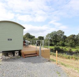 An outdoor area with a small green building wooden deck patio furniture and a river with trees in the background at Llety'r Bugail 1 in Llanrug