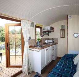 A compact kitchen with a sink microwave and toaster next to a blue patterned sofa and an open door leading outside at Llety'r Bugail 1 in Llanrug