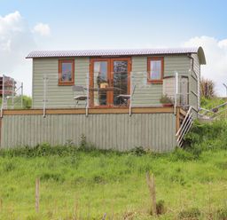 A small green cabin with glass railings and outdoor furniture on a raised platform with stairs surrounded by grass at Llety'r Bugail 1 in Llanrug