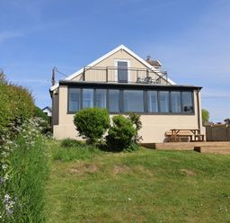 The back of a two-story house with a balcony and enclosed porch overlooking a grassy yard with bushes and a wooden picnic table on a deck at Tillicum in Charmouth